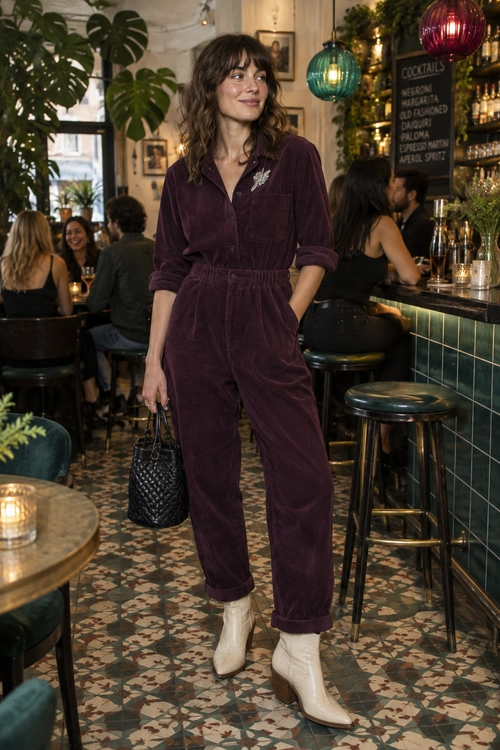 Plum Velvet Jumpsuit Outfit A plum velvet button-front jumpsuit with a floral brooch, black woven handbag, and cream ankle boots.